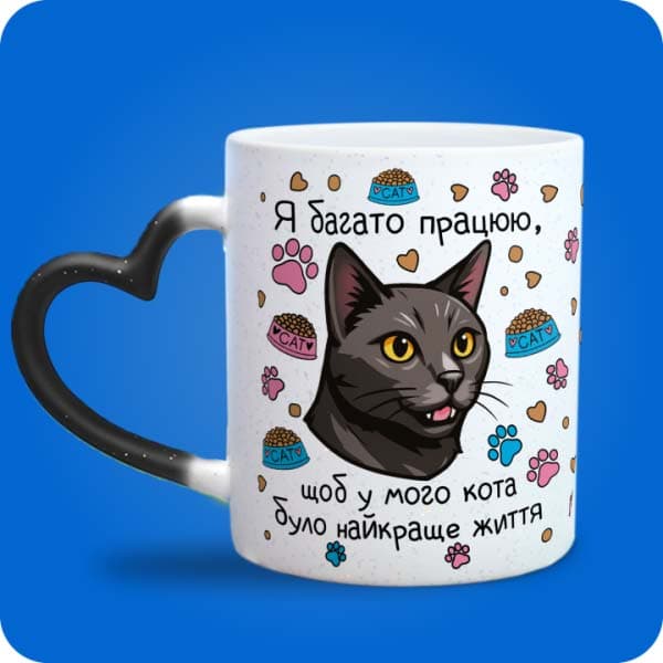 Біла чашка з чорним котом, лапками, кормом і написом "Я багато працюю, щоб у мого кота було найкраще життя", вигляд чашки хамелеон з ручкою сердечко
